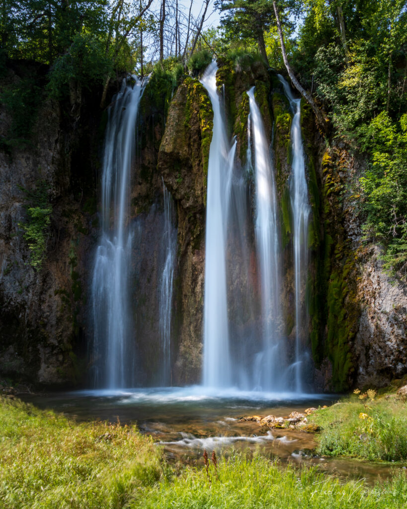 Spearfish Falls cascades down a rugged rock face into a serene pool, surrounded by lush greenery in Spearfish Canyon, South Dakota.