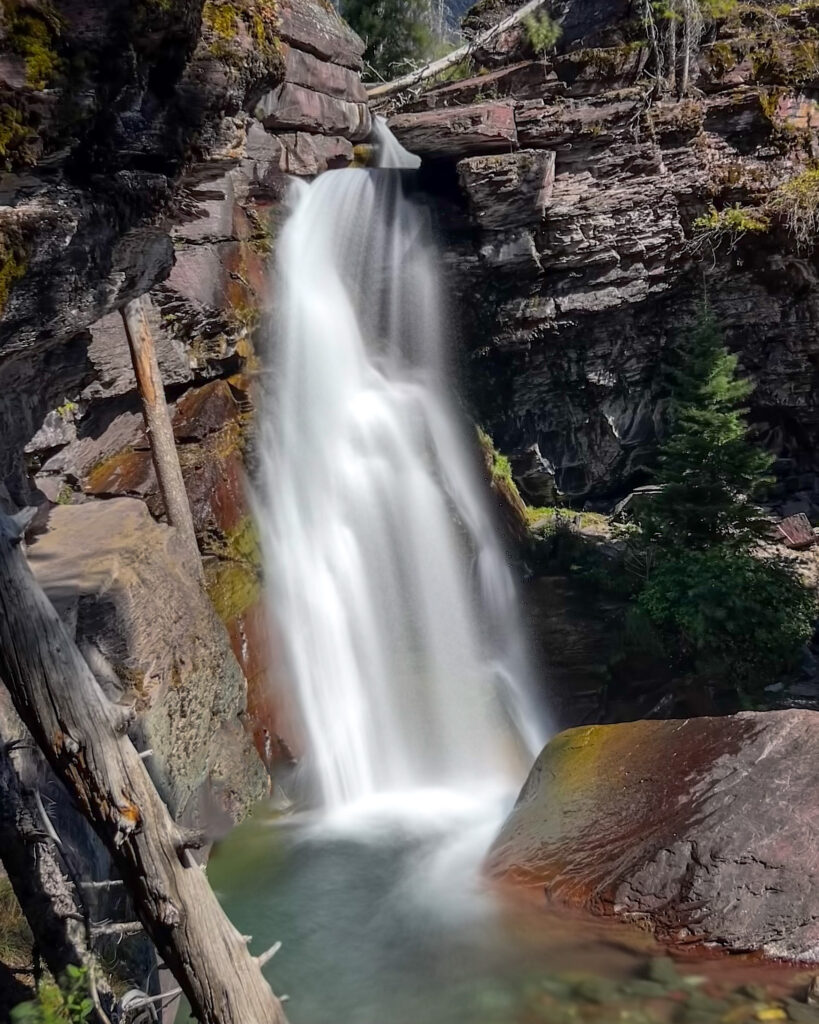 A stunning waterfall cascades down rocky cliffs in Glacier National Park, surrounded by lush greenery. The water tumbles over jagged rocks, creating a misty spray that glistens in the sunlight.