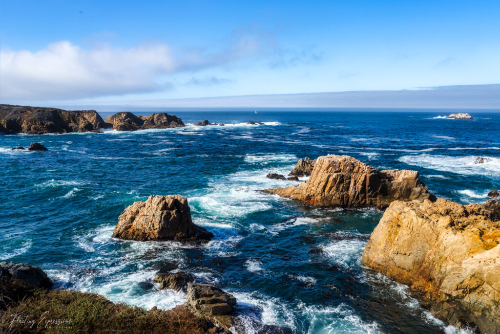 View from Painter’s Point along California Highway 1 in Big Sur, showcasing the rugged coastline with cliffs dropping into the Pacific Ocean. The sky is a bright blue with a few white clouds scattered above, adding to the scenic beauty.